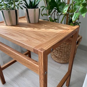 May include: A rectangular side table constructed from light brown wood with a visible grain pattern. The table features four straight legs connected by horizontal support bars and is assembled with visible silver-toned metal circular fasteners.