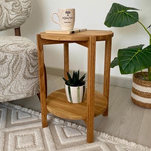 May include: A round wooden side table with a natural light brown finish, featuring three vertical legs and a lower shelf. The table is constructed from solid wood with a visible grain pattern, providing a stable surface for a ceramic mug and a small plant.