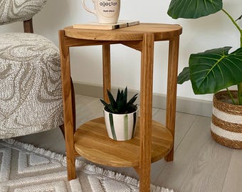 May include: A round wooden side table with a natural light brown finish, featuring three vertical legs and a lower shelf. The table is constructed from solid wood with a visible grain pattern, providing a stable surface for a ceramic mug and a small plant.
