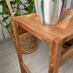 May include: A light brown wooden side table with a rectangular top and four legs, featuring visible silver metal circular fasteners at each corner joint. A ceramic plant pot with vertical green and white stripes sits on the table surface.