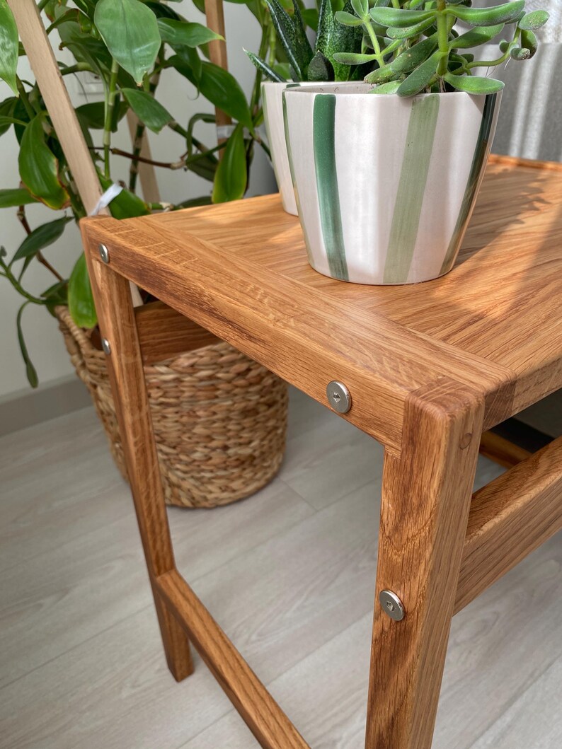 May include: A light brown wooden side table with a rectangular top and four legs, featuring visible silver metal circular fasteners at each corner joint. A ceramic plant pot with vertical green and white stripes sits on the table surface.
