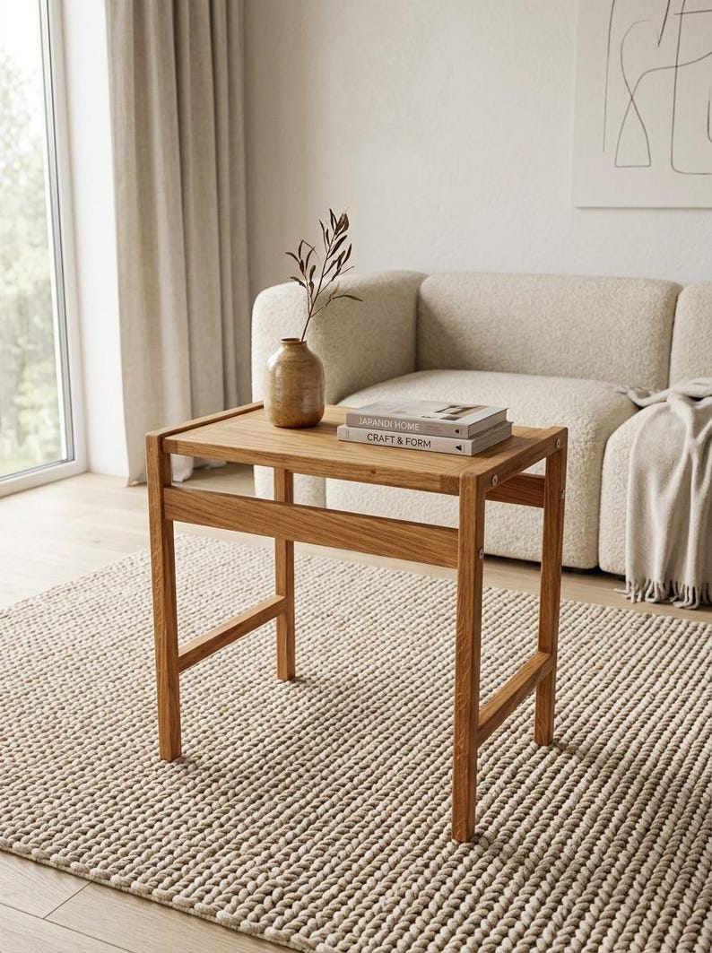 May include: A rectangular wooden side table with four straight legs and a horizontal support bar. On the tabletop sits a tan ceramic vase containing a single dried branch and two stacked books titled Japandi Home and Craft & Form.