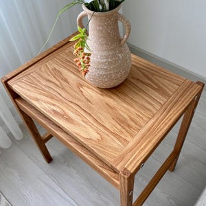 May include: A rectangular wooden side table with a natural grain finish and four straight legs, featuring a light beige speckled ceramic vase with two handles placed on top, containing a bouquet of white flowers and green foliage.