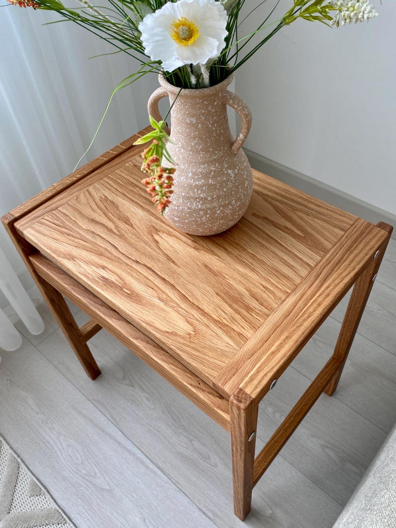 May include: A rectangular wooden side table with a natural grain finish and four straight legs, featuring a light beige speckled ceramic vase with two handles placed on top, containing a bouquet of white flowers and green foliage.