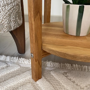 K&ouml;nnte beinhalten: Ein runder Beistelltisch aus Holz mit nat&uuml;rlichem Eichenfinish, bestehend aus einer runden Tischplatte und vier geraden Beinen, die durch eine untere Ablage verbunden sind. Eine kleine silberne Metallschraube ist am Bein sichtbar. Ein gr&uuml;n gestreifter Topf.