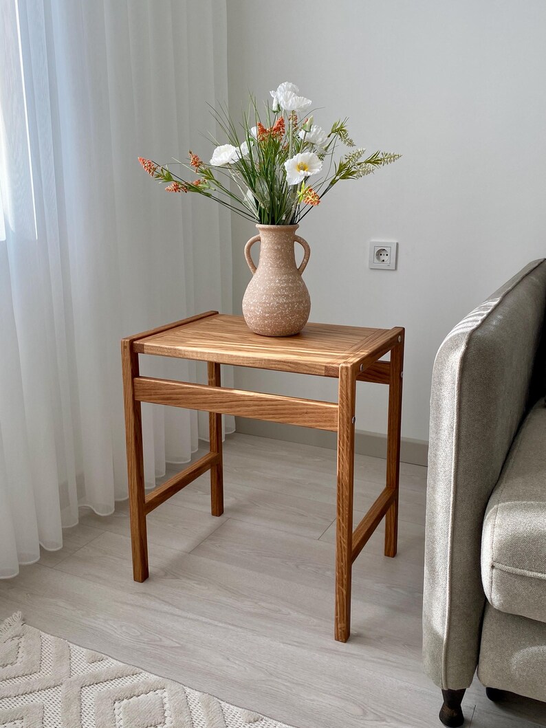 May include: A small, wooden side table with a vase of flowers. The table is made of light-colored wood and has a rectangular top. The vase is a light brown color and has two handles. The flowers are white and orange.