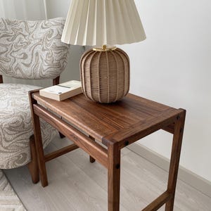 May include: A wooden side table with a woven lamp and a book. The lamp has a beige pleated shade and a brown woven base. The table is made of dark wood and has a rectangular top. A patterned chair is in the background.