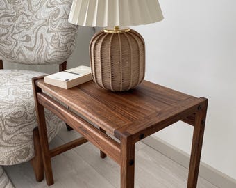 Könnte beinhalten: Ein Beistelltisch aus Holz mit einer gewebten Lampe und einem Buch. Die Lampe hat einen beigefarbenen plissierten Schirm und einen braunen gewebten Sockel. Der Tisch ist aus dunklem Holz gefertigt und hat eine rechteckige Platte. Ein Buch liegt auf dem Tisch.
