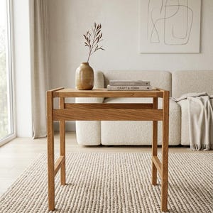 May include: A light brown wooden console table with a rectangular top and four straight legs. On the table surface sits a tan ceramic vase containing a single dried branch and two stacked books titled Japandi Home and Craft & Form.