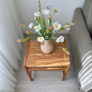 May include: A square wooden side table with a light brown grain pattern, topped with a speckled beige ceramic vase containing an arrangement of white poppy-like flowers, yellow centers, green stems, and small orange-tipped floral accents.