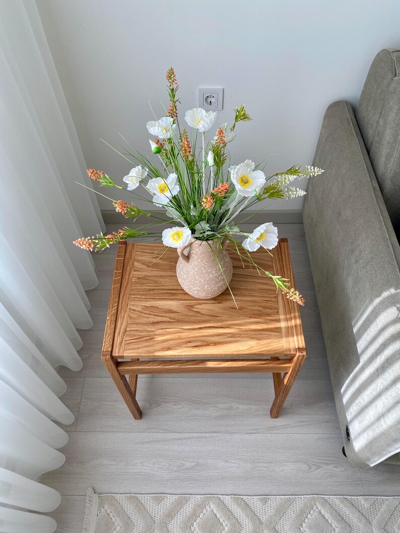 May include: A square wooden side table with a light brown grain pattern, topped with a speckled beige ceramic vase containing an arrangement of white poppy-like flowers, yellow centers, green stems, and small orange-tipped floral accents.