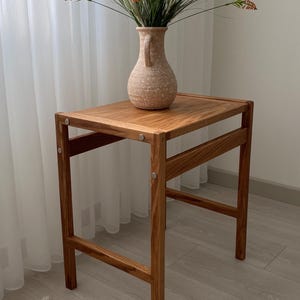 May include: A rectangular wooden side table with a natural grain finish and visible metal hardware connecting the frame. A speckled beige ceramic vase with a single handle sits on the tabletop, containing a bouquet of white and orange flowers.