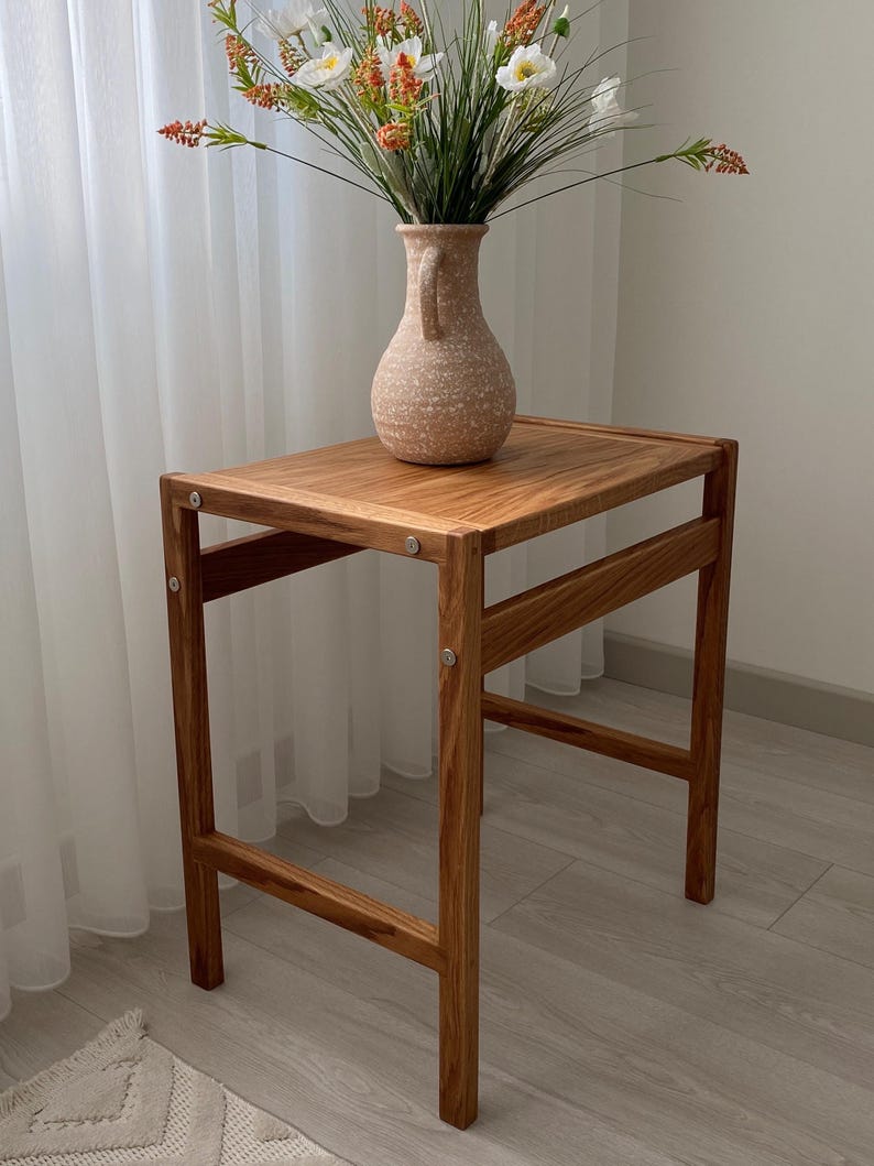 May include: A rectangular wooden side table with a natural grain finish and visible metal hardware connecting the frame. A speckled beige ceramic vase with a single handle sits on the tabletop, containing a bouquet of white and orange flowers.