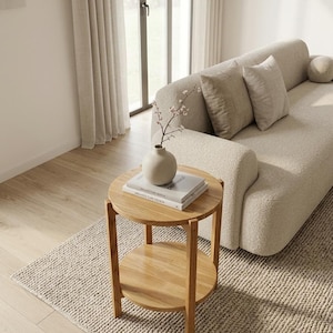 K&ouml;nnte beinhalten: Ein runder, zweist&ouml;ckiger Beistelltisch aus Holz mit hellbraunem Finish. Der Tisch verf&uuml;gt &uuml;ber vier gerade Beine und eine runde untere Ablage. Eine matte, beige Keramikvase mit einem zarten Zweig steht auf zwei gestapelten B&uuml;chern.