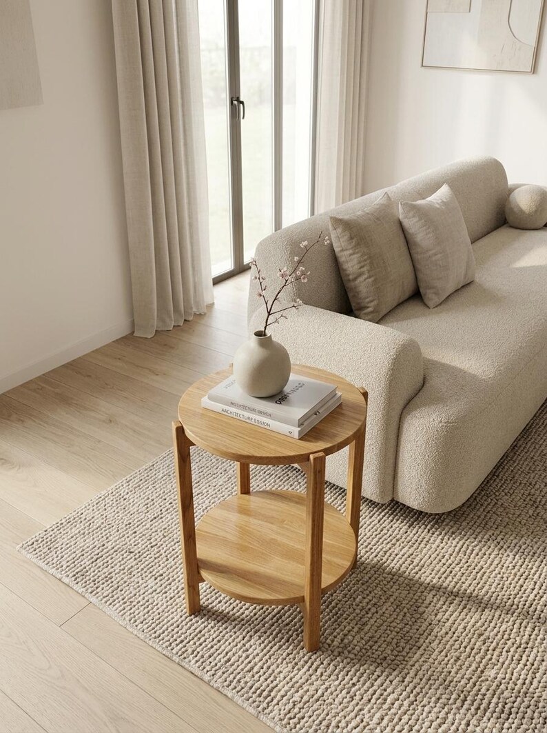 K&ouml;nnte beinhalten: Ein runder, zweist&ouml;ckiger Beistelltisch aus Holz mit hellbraunem Finish. Der Tisch verf&uuml;gt &uuml;ber vier gerade Beine und eine runde untere Ablage. Eine matte, beige Keramikvase mit einem zarten Zweig steht auf zwei gestapelten B&uuml;chern.