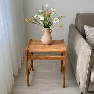May include: A square wooden side table with a natural grain finish and four straight legs, topped with a light beige speckled ceramic vase featuring two side handles and a bouquet of white and orange artificial flowers with green foliage.