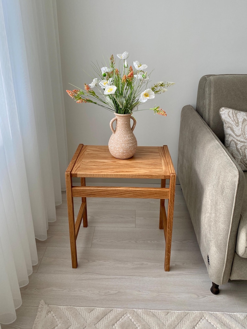 May include: A square wooden side table with a natural grain finish and four straight legs, topped with a light beige speckled ceramic vase featuring two side handles and a bouquet of white and orange artificial flowers with green foliage.