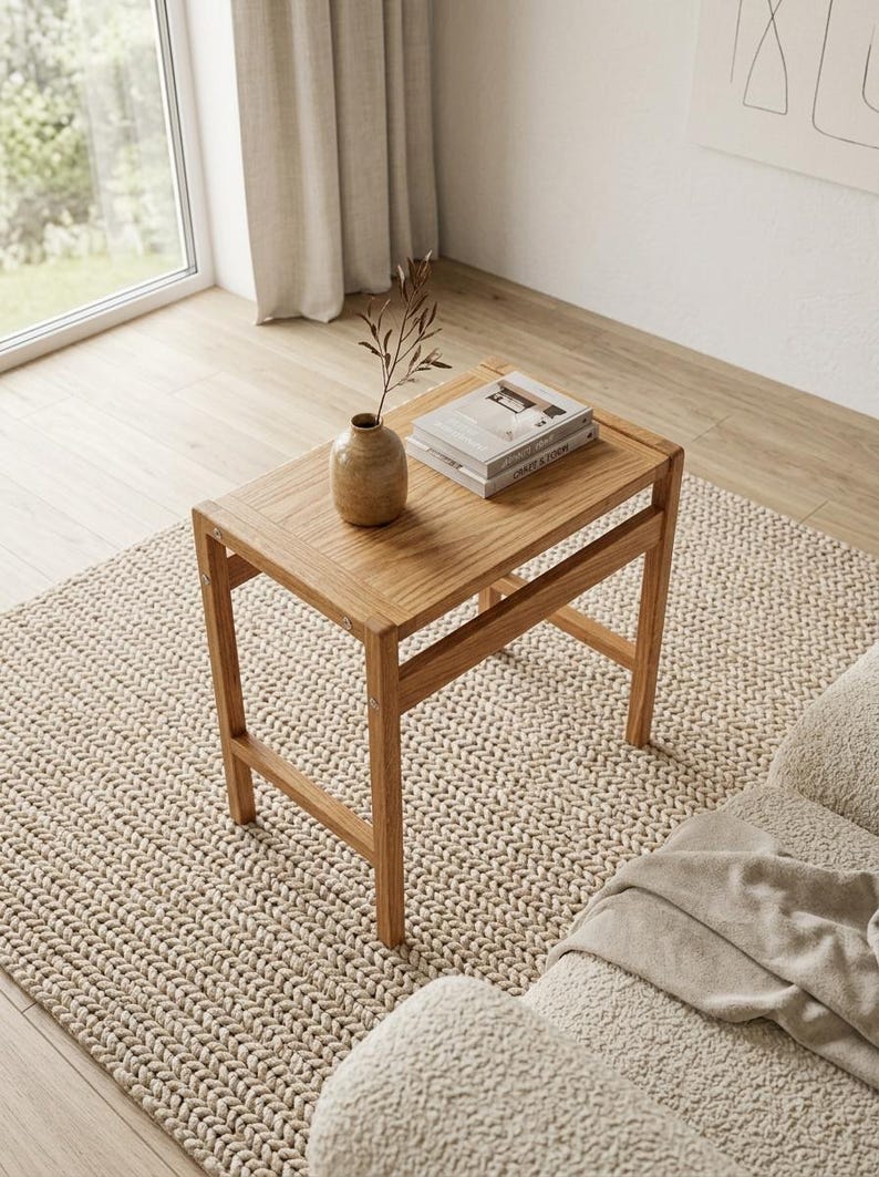 May include: A rectangular light brown wooden side table with four legs and a slatted top. On the table sits a small tan ceramic vase containing a single dried branch and a stack of two books with white covers and visible text on the spines.