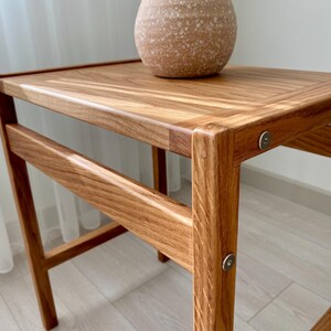 May include: A wooden side table with a rectangular top and four legs, featuring visible metal circular fasteners at the joints. A speckled, light tan ceramic vase with two handles sits on the surface of the table, which has a natural wood grain finish.