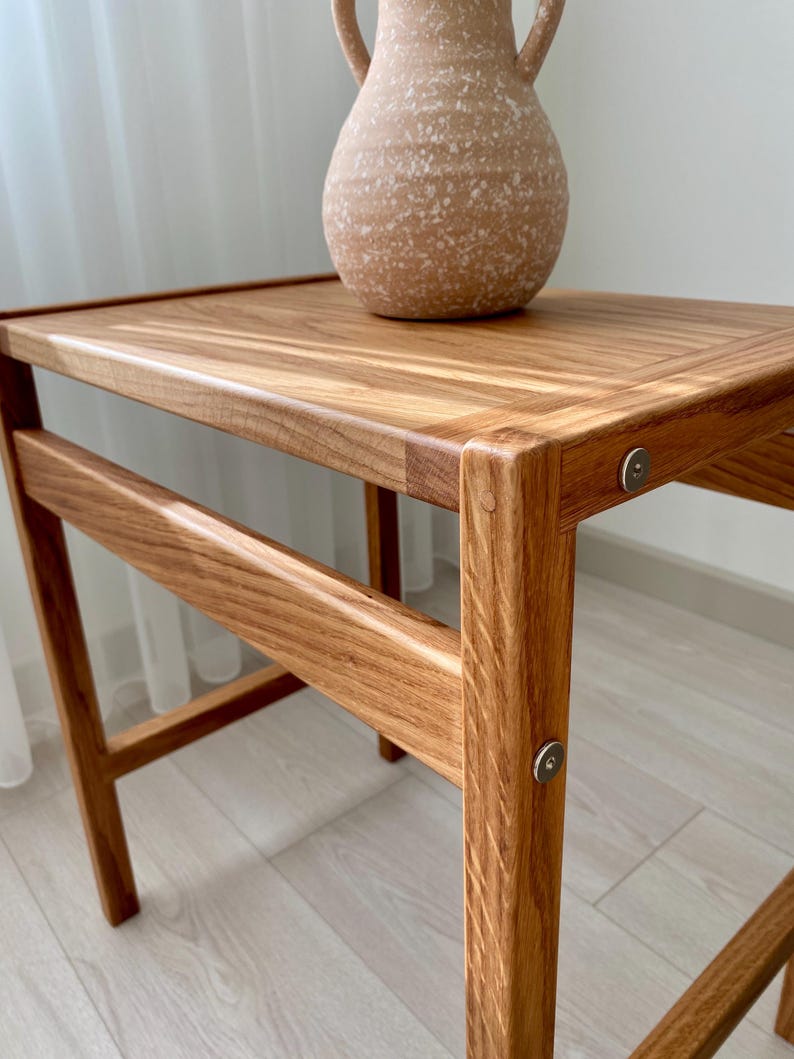 May include: A wooden side table with a rectangular top and four legs, featuring visible metal circular fasteners at the joints. A speckled, light tan ceramic vase with two handles sits on the surface of the table, which has a natural wood grain finish.