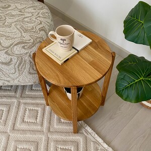 K&ouml;nnte beinhalten: Ein runder, zweist&ouml;ckiger Beistelltisch aus Holz mit einer nat&uuml;rlichen hellbraunen Oberfl&auml;che. Auf der oberen Ablage steht eine cremefarbene Keramiktasse mit einem Baum-Grafikmotiv und dem Text "ağa&ccedil;tan" sowie ein Buch mit dem Titel "Victor Hugo".