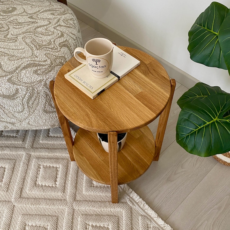 K&ouml;nnte beinhalten: Ein runder, zweist&ouml;ckiger Beistelltisch aus Holz mit einer nat&uuml;rlichen hellbraunen Oberfl&auml;che. Auf der oberen Ablage steht eine cremefarbene Keramiktasse mit einem Baum-Grafikmotiv und dem Text "ağa&ccedil;tan" sowie ein Buch mit dem Titel "Victor Hugo".