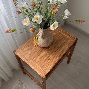May include: A light brown wooden side table with a rectangular top and four legs, featuring a speckled beige ceramic vase filled with white poppy-like flowers and orange-tipped green stalks arranged in a decorative floral display.