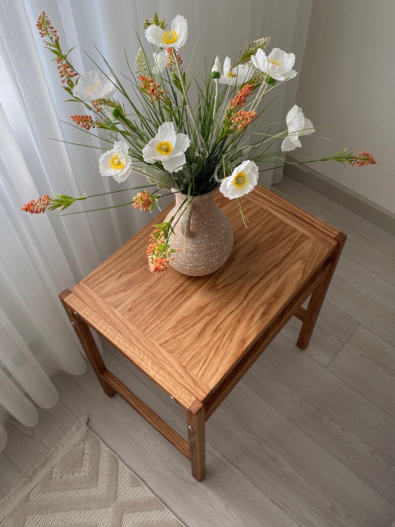 May include: A light brown wooden side table with a rectangular top and four legs, featuring a speckled beige ceramic vase filled with white poppy-like flowers and orange-tipped green stalks arranged in a decorative floral display.