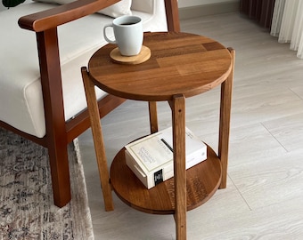 May include: A round wooden side table with two tiers, featuring a natural brown wood grain finish. A white ceramic mug sits on a small wooden coaster on the top surface, while a white book rests on the lower shelf of the table structure.