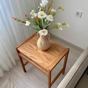 May include: A rectangular wooden side table with a natural grain finish and four legs, topped with a speckled beige ceramic vase featuring two handles. The vase contains an arrangement of white poppy flowers, green grass, and small orange floral sprigs.