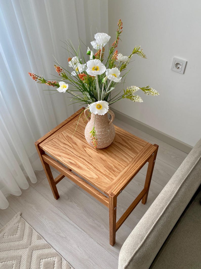 May include: A rectangular wooden side table with a natural grain finish and four legs, topped with a speckled beige ceramic vase featuring two handles. The vase contains an arrangement of white poppy flowers, green grass, and small orange floral sprigs.