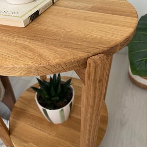 K&ouml;nnte beinhalten: Ein runder Beistelltisch aus hellem Holz mit sichtbarer Maserung. Der Tisch verf&uuml;gt &uuml;ber eine kreisf&ouml;rmige Tischplatte und eine untere Ablage, die von vertikalen Holzbeinen gest&uuml;tzt wird. Ein Buch und eine kleine Sukkulente stehen darauf.