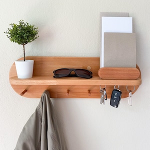 May include: A wooden wall-mounted organizer with a shelf, key hooks, and a small plant in a white pot. The shelf holds sunglasses and a small tray for mail. A jacket hangs from one of the hooks. The organizer is made of light-colored wood.