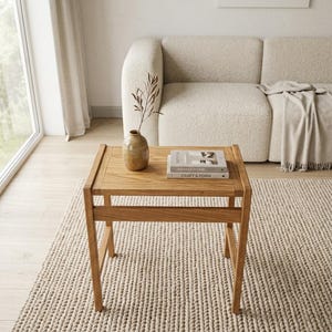 May include: A square wooden side table with a light brown finish, featuring four straight legs and a horizontal support frame. On the tabletop sits a small, speckled tan ceramic vase containing a single dried branch and two stacked books.