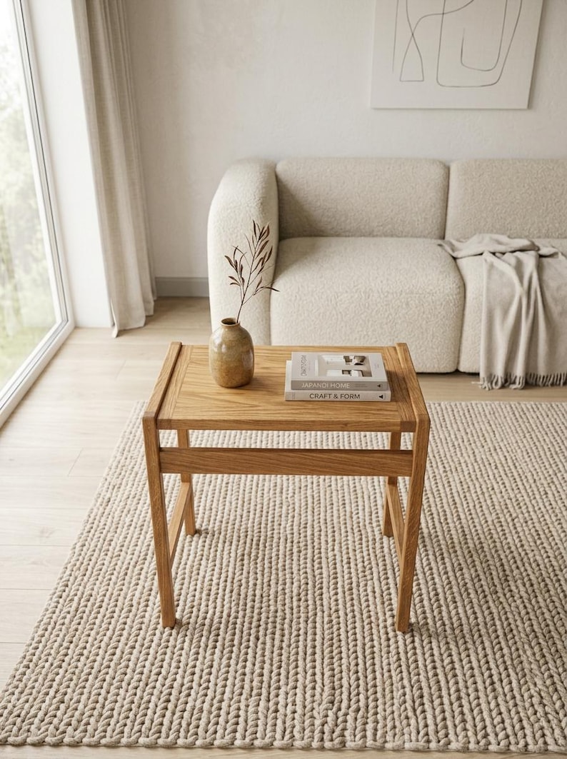 May include: A square wooden side table with a light brown finish, featuring four straight legs and a horizontal support frame. On the tabletop sits a small, speckled tan ceramic vase containing a single dried branch and two stacked books.