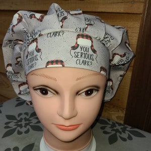 May include: A scrub cap featuring a light gray background with a repeating pattern of red and black buffalo plaid trapper hats and the text "YOU SERIOUS CLARK?" in black lettering. The cap has a pleated design and a wide, structured front band.