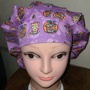 May include: Purple scrub hat with a Halloween theme. The hat has a pattern of pumpkins, bats, and ghosts. The text "TRACH OR TREAT!" is printed on the hat.