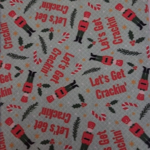 May include: A light grey fabric featuring a repeating pattern of red and green nutcracker figures, red candy canes, green pine sprigs, and holly berries. The text Let's Get Crackin' is printed in red throughout the design in a repeating layout.