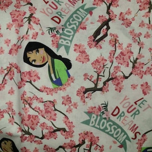 May include: A white fabric with a pink floral pattern and a cartoon image of a woman with black hair. The fabric has the text "Let your dreams blossom" repeated on it.