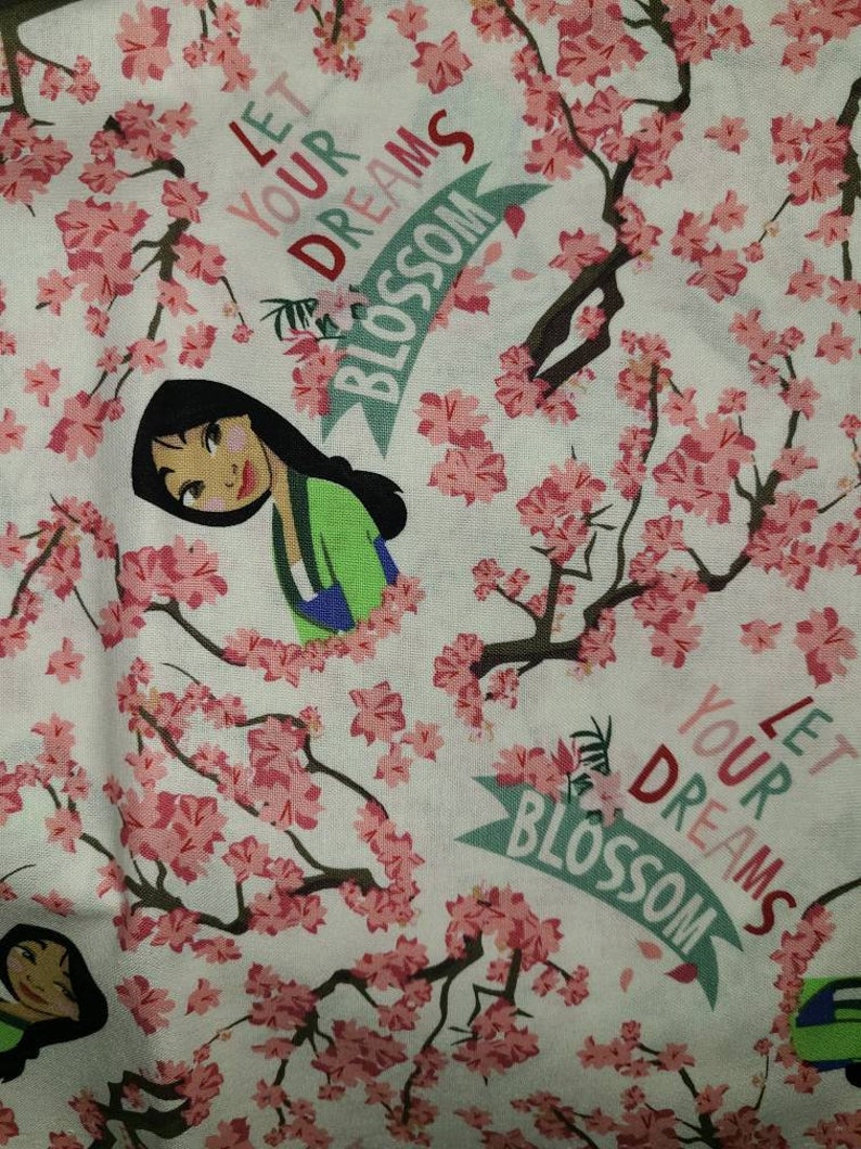 May include: A white fabric with a pink floral pattern and a cartoon image of a woman with black hair. The fabric has the text "Let your dreams blossom" repeated on it.