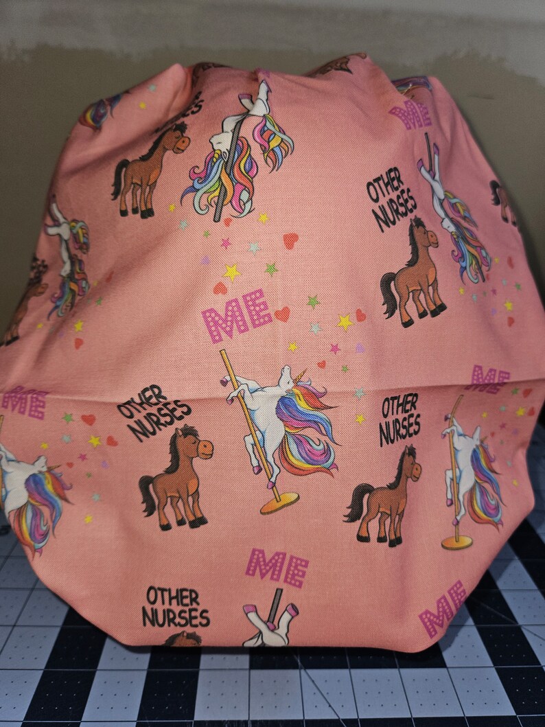 K&ouml;nnte beinhalten: Rosafarbener Stoff mit einem sich wiederholenden Muster aus Einh&ouml;rnern auf einer Stange, Pferden und dem Text "Other Nurses" und "Me".