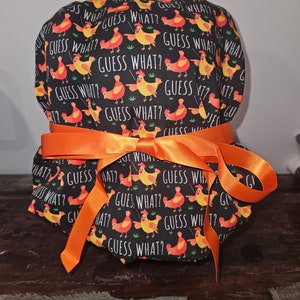 May include: A black fabric scrub cap with a repeating pattern of orange and yellow chickens and the text "GUESS WHAT?". The cap is tied with a bright orange ribbon.