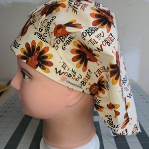 May include: A white scrub hat with a turkey print and the text "Gobble Til You Wobble!" in orange and brown. The hat has a tie on the back.