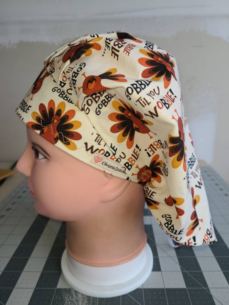 May include: A white scrub hat with a turkey print and the text "Gobble Til You Wobble!" in orange and brown. The hat has a tie on the back.