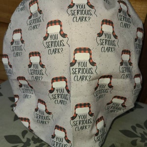May include: A scrub cap featuring a light gray fabric with a repeating pattern of red and black buffalo plaid trapper hats and the text "YOU SERIOUS CLARK?" printed in black. The cap has a rounded, gathered shape with a finished hem at the bottom.