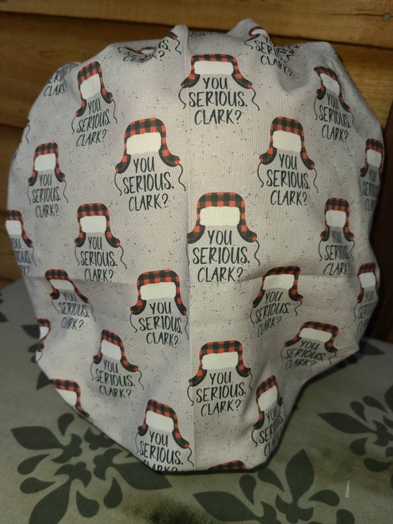 May include: A scrub cap featuring a light gray fabric with a repeating pattern of red and black buffalo plaid trapper hats and the text "YOU SERIOUS CLARK?" printed in black. The cap has a rounded, gathered shape with a finished hem at the bottom.