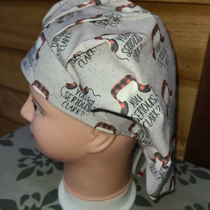 May include: A light gray scrub cap featuring a repeating pattern of black and red buffalo plaid trapper hats and the text "YOU SERIOUS CLARK?" in black lettering. The fabric has a subtle speckled texture throughout the design.