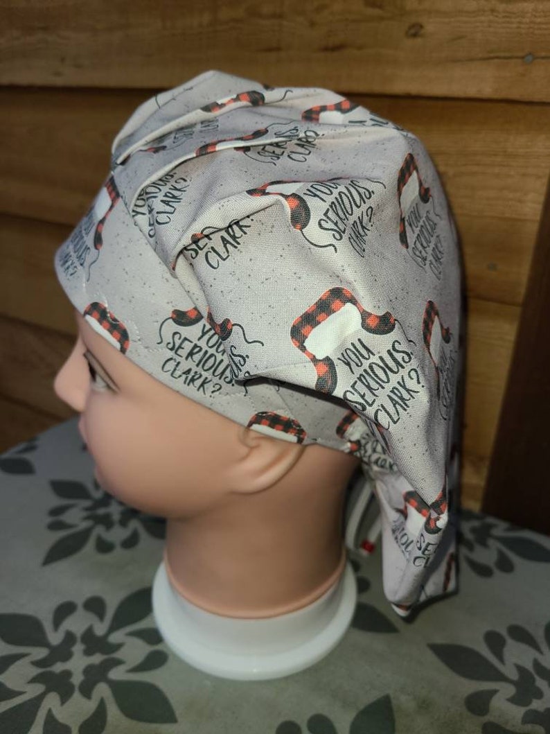 May include: A light gray scrub cap featuring a repeating pattern of black and red buffalo plaid trapper hats and the text "YOU SERIOUS CLARK?" in black lettering. The fabric has a subtle speckled texture throughout the design.