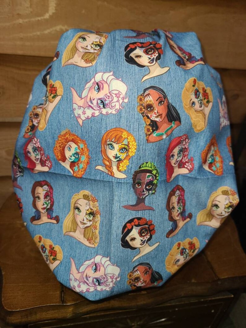 May include: A blue fabric with a pattern of various Disney princesses with sugar skull makeup. The princesses include Elsa, Anna, Belle, Ariel, Snow White, and Rapunzel.