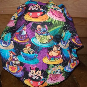 May include: A colorful fabric scrub hat with a black background and a galaxy print. The hat features various Disney characters riding in teacups, including Mickey Mouse, Minnie Mouse, Stitch, and Woody.